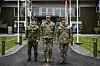 Tre uniformerte soldater står på en gangvei foran et bygg med flere flagg.