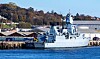 Et grått skip ligger ved kai foran lagerbygninger og en havn med trær i bakgrunnen.