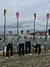 Fire roere står på en brygge med årer mot en by og vann i bakgrunnen.