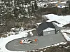 Helikopter på en rund landingsplass ved en grå bygning i et snødekt skoglandskap.