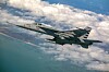 Militært jagerfly i luften over skyer, hav og kystlinje.
