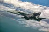 Militært jagerfly i luften over skyer, hav og kystlinje.