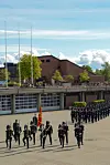 Uniformerte personer marsjerer i formasjon foran et teglbygg ved vannet.