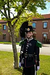 Vakt i mørk seremoniell uniform står under et tre foran en murbygning.